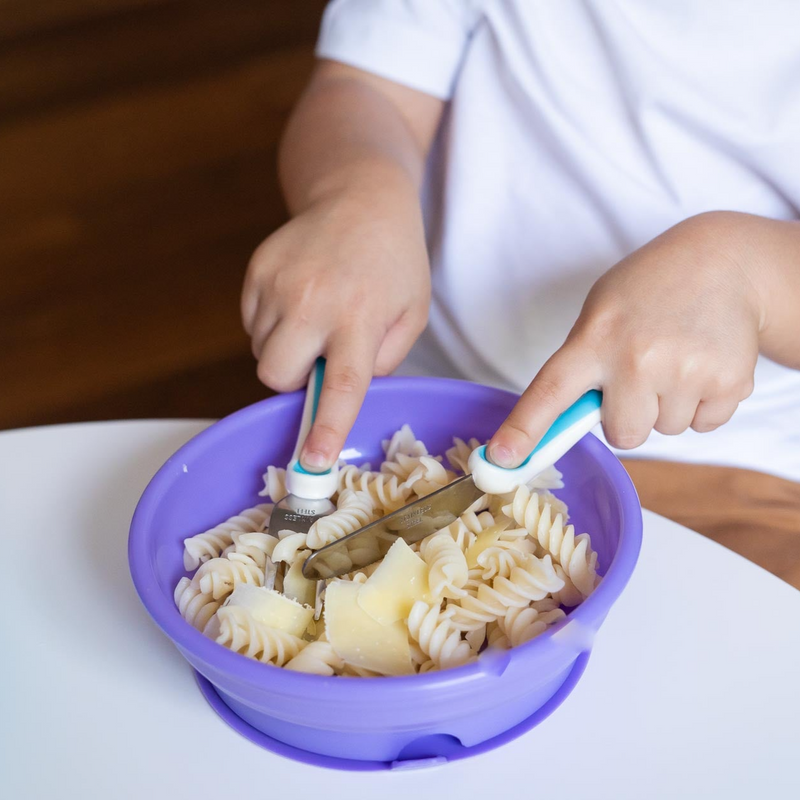 Ergonomic children’s training cutlery set in blue being used with toddler bowl by young child 